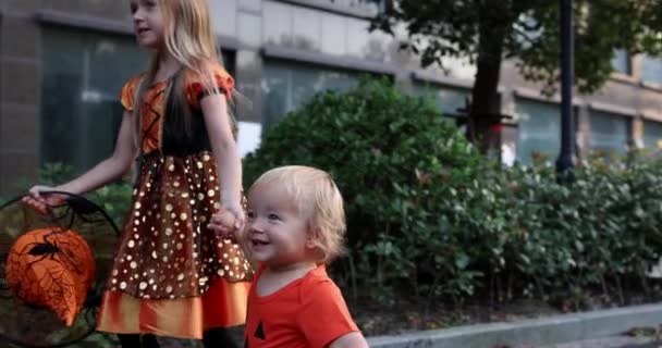 Söta små kaukasiska barn med blont hår sju år gammal i dräkt av häxa med hatt och baby ett år gammal firar Halloween utomhus på gatan. Semesterkoncept. Långsamma rörelser. — Stockvideo