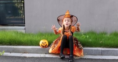 Yedi yaşında, sarı saçlı, şapkalı, siyah turuncu elbiseli, caddede cadılar bayramını kutlayan bir cadı kostümü giyen sevimli, beyaz bir kız. Tatil konsepti. Yavaş çekim.