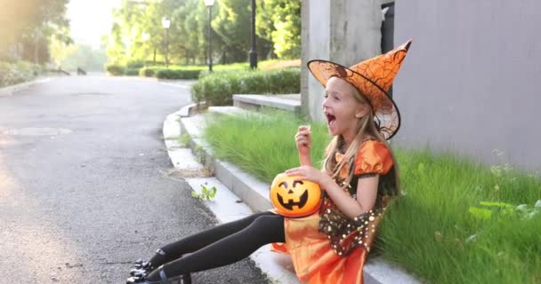 Schattig klein blank meisje met blond haar zeven jaar oud in kostuum van heks met hoed en zwarte oranje jurk vieren Halloween buiten op straat. Vakantie concept. Langzame beweging. — Stockvideo