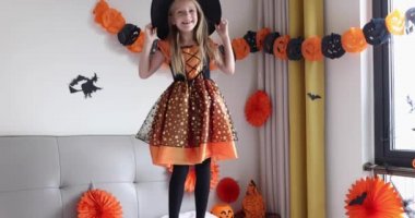 Cadı elbisesi giymiş, şapkalı, sarı saçlı sevimli beyaz kız Coronavirus covid-19 salgını ve karantinası sırasında evde eğleniyor ve Cadılar Bayramı 'nı kutluyor. Yavaş çekim