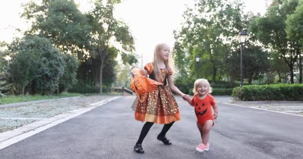 Leuke kleine blanke kinderen met blond haar zeven jaar oud in kostuum van heks met hoed en baby een jaar oud vieren Halloween buiten op straat. Vakantie concept. Langzame beweging. — Stockvideo