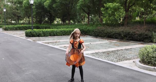 Söt liten kaukasiska flicka med blont hår sju år gammal i dräkt av häxa med hatt och svart orange klänning poserar med gäng halloween ballonger på gatan. Semesterkoncept. Långsamma rörelser. — Stockvideo