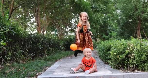 Leuke kleine blanke kinderen met blond haar zeven jaar oud in kostuum van heks met hoed en baby een jaar oud vieren Halloween buiten op straat. Vakantie concept. Langzame beweging. — Stockvideo