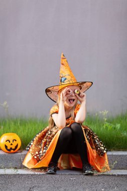 Sarı saçlı, siyah turuncu kostümlü küçük mutlu beyaz kızın yaşam tarzı portresi. Coronavirus covid-19 pandemik karantinası sırasında dışarıda tek başına Cadılar Bayramı 'nı kutluyor..