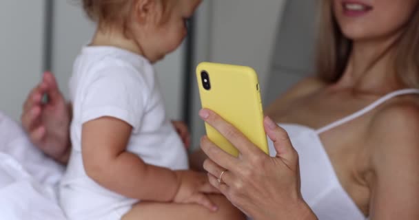Молодая кавказская мать, пользующаяся мобильным телефоном и работающая дома с дочкой годовалого возраста. Счастливая семейная мама и милый маленький сын учатся веселой игре, получая удовольствие от сладких моментов — стоковое видео