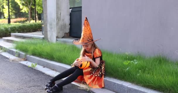 Roztomilý malý kavkazský dívka s blond vlasy sedm let v kostýmu čarodějnice s kloboukem a černooranžové šaty slaví Halloween venkovní na ulici. Koncept dovolené. Zpomalený pohyb. — Stock video