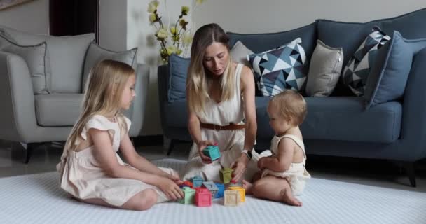 Young caucasian mother or nanny playing with cute baby one year old and elder daughter, learning to build a tower from cubes for kids. Three generation family spending time together and enjoying life — Stock Video