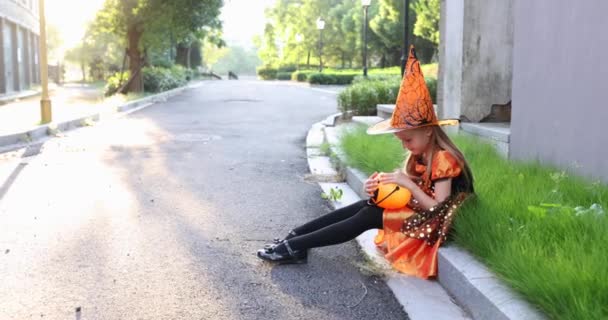 Schattig klein blank meisje met blond haar zeven jaar oud in kostuum van heks met hoed en zwarte oranje jurk vieren Halloween buiten op straat. Vakantie concept. Langzame beweging. — Stockvideo