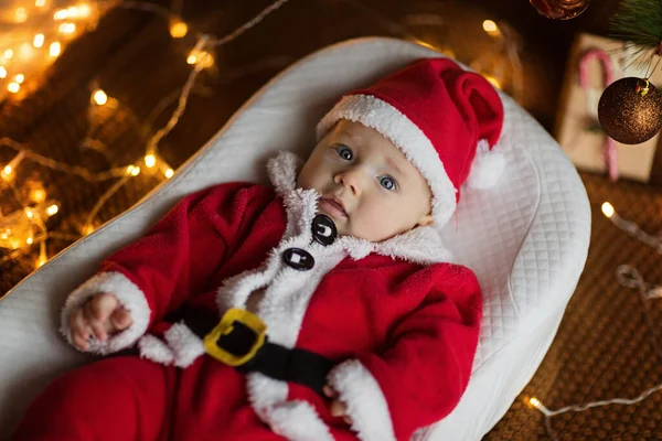 Portrait rapproché du nouveau-né. Mignon bébé fille blanche 4-5 mois en costume de Père Noël couché sur une couverture confortable tricotée dans un cocon près d'un sapin décoré. Joyeux Noël et bonne année 2022 — Photo