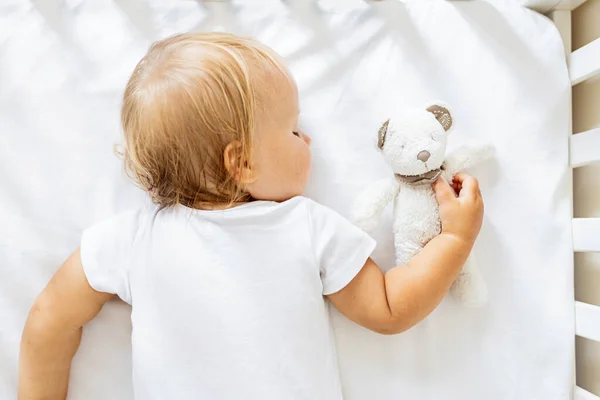 Schattige kleine baby van een jaar oud met blond haar knuffelende knuffelende knuffelbeer speelgoed wanneer liggend op witte lakens in gezellige slaapkamer thuis dutje dagdroom. Klein kind slaapt op bed, droomt of rust alleen — Stockfoto