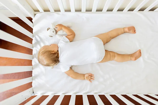 Schattige kleine baby van een jaar oud met blond haar knuffelende knuffelende knuffelbeer speelgoed wanneer liggend op witte lakens in gezellige slaapkamer thuis dutje dagdroom. Klein kind slaapt op bed, droomt of rust alleen — Stockfoto