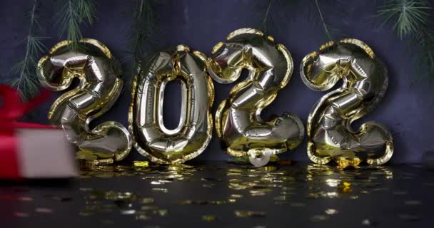 Frohes neues Jahr 2022 Hintergrund. Metallische Luftballons goldene Farbe, umhüllte Geschenkbox auf grauem Beton mit Konfetti. Winterfest. Zeitlupe — Stockvideo