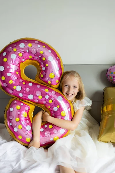 Vnitřní záběr docela radostné roztomilé roztomilé holčičky oslavující osm let staré narozeniny s jasnými a barevnými balónky s nápisem slova zůstat báječný, na sobě ležérní módní šaty — Stock fotografie