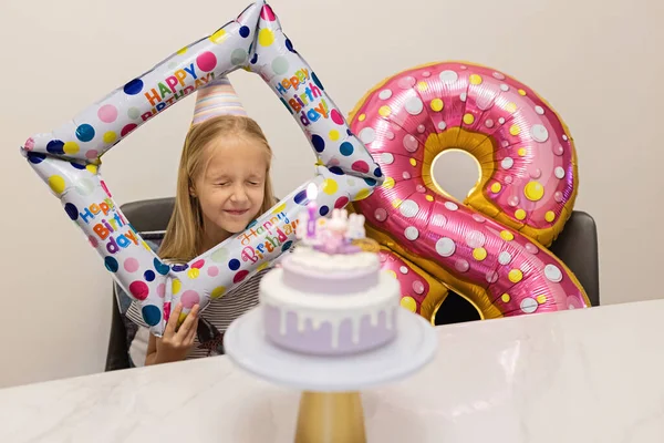 Boldog kis kaukázusi lány pozitív érzelmekkel az arcán, aki 8 éves születésnapját ünnepli otthon. Őszinte életmód portré Kölyökről, amint gyertyákat fúj a tortára. Nappali fóliával díszítve — Stock Fotó