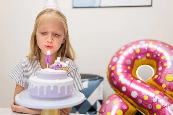 Glückliches kleines kaukasisches Mädchen mit positiver Emotion im Gesicht, das zu Hause seinen achten Geburtstag feiert. Ehrliches Lifestyleporträt von Kid, das Kerzen auf Kuchen pustet. Wohnzimmer mit Folie dekoriert — Stockfoto