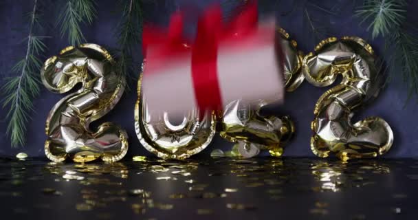 Gott nytt år 2022 bakgrund. Metalliska ballonger gyllene färg, insvept presentförpackning på grå betong med konfetti. Vintersemester firande. Långsamma rörelser — Stockvideo