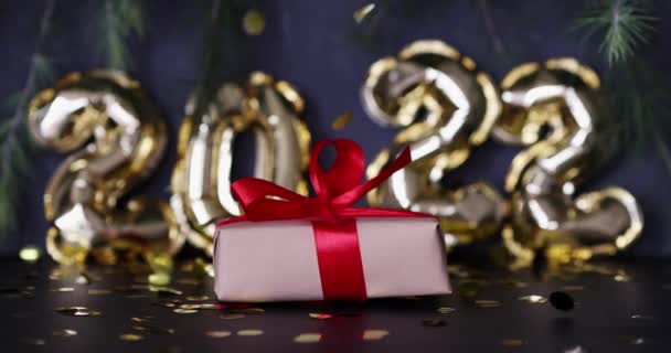 Boldog új évet 2022 háttér. Fémes lufik arany színű, becsomagolt ajándékdoboz szürke betonon konfettivel. Téli ünnepség. Lassú mozgás. — Stock videók