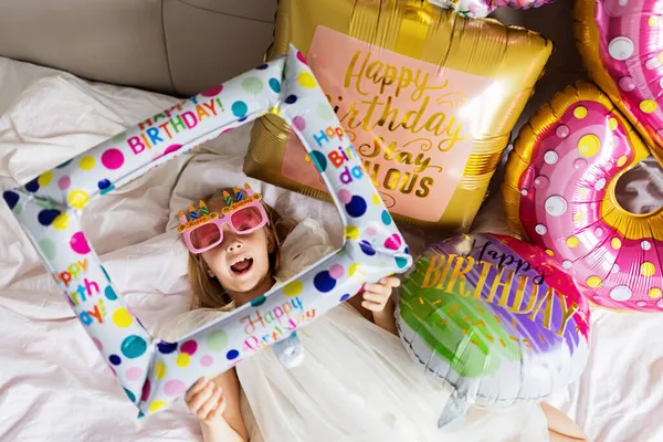 Indoor shot of pretty joyful cute adorable girl kid celebrando el cumpleaños de ocho años con globos brillantes y coloridos con palabras de inscripción permanecen fabulosas, vistiendo vestido casual de moda — Foto de Stock