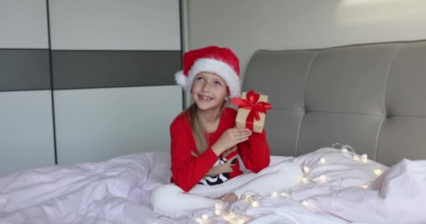 Feliz adorable niña caucásica con el pelo rubio de ocho años de edad con sombrero de Santa rojo y suéter de punto con caja de regalo en la cama en casa. Niño celebrando la Navidad. Feliz año nuevo 2022 — Vídeo de stock