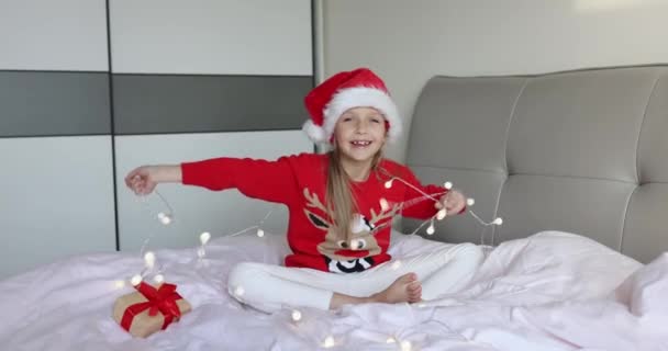 Счастливая очаровательная кавказская маленькая девочка с светлыми волосами восьми лет в красной шляпе Санты и вязаном свитере, когда весело проводишь время дома с гирляндой. Ребёнок празднует Рождество. С новым 2022 годом! — стоковое видео