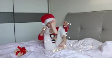Sekiz yaşında, sarı saçlı, kırmızı Noel Baba şapkası takan ve evde çelenkle eğlenirken örgü ören sevimli beyaz kız. Noel 'i kutlayan çocuk. Mutlu yıllar 2022