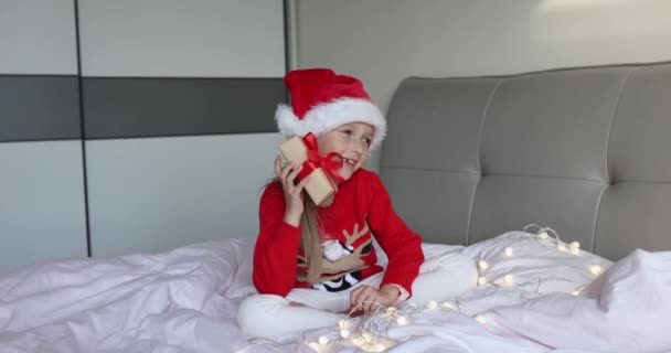 Menina caucasiana adorável feliz com cabelo loiro oito anos de idade vestindo chapéu de Papai Noel vermelho e camisola de malha com caixa de presente na cama em casa. Criança a celebrar o Natal. Feliz ano novo 2022 — Vídeo de Stock