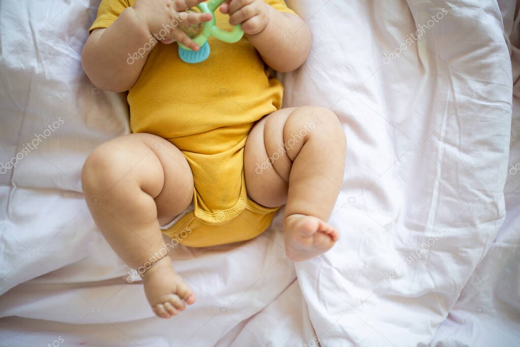 Caucasian blonde baby seven months old lying on bed at home. Kid wearing cute clothing trendy colors: illuminating yellow. Infant playing with toys