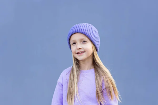 Χαριτωμένο μικρό καυκάσιο κορίτσι οκτώ ετών με ξανθά μαλλιά χαμογελαστά εξωτερικά. Παιδί που φοράει κομψό πουκάμισο και πλεκτό καπέλο βιολετί χρώμα. Trendy χρώμα του έτους 2022 πολύ peri — Φωτογραφία Αρχείου
