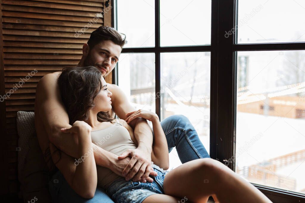 portrait of a charming young couple at home. man is embracing his girlfriend near window. Two people relaxing together