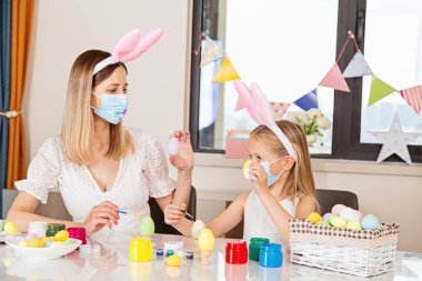 Tavşan kulağı takan, tek kullanımlık tıbbi maske takan ve Coronavirus covid-19 salgını sırasında evde paskalya yumurtası boyayan güzel anne ve çocuk.. 