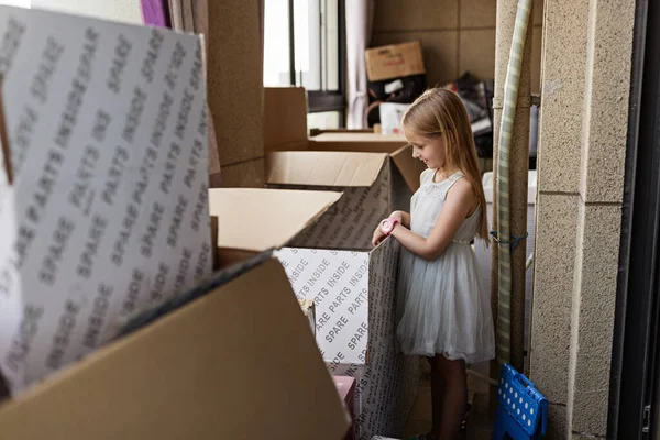 Lifestyle portrait of happy little caucasian girl six years old with blonde hair playing at home with cardboard boxes in new house at moving day. — Stok fotoğraf