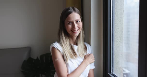 Glückliche junge kaukasische Frau mit blonden Haaren zeigt COVID-19 Impfverband am Arm, wenn sie zu Hause am Fenster steht — Stockvideo
