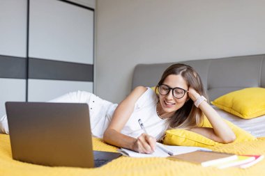Young Woman student studying online via video chat. Stylish freelancer works from home. Blogger working from home office. Millennial business woman lying on bed in modern apartment. 