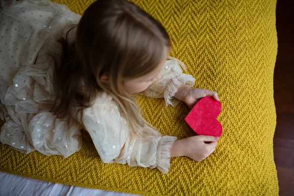 Een schoolmeisje met een rood hart. Valentijnsdag tijdens pandemische coronavirus covid-19 quarantaine. Kind tonen hart online via video chat naar jongen vriend — Stockfoto