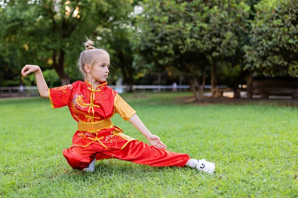 Cute little caucasian girl seven years old in red sport wushu uniform exercising in park at summer day. Lifestyle portrait of kung fu fighter child athlete — Stock Photo, Image