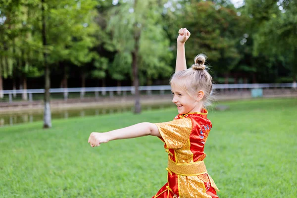 Cute little kaukaska dziewczyna siedem lat w czerwonym sporcie wushu mundur ćwiczenia w parku w letni dzień. Portret stylu życia wojownika kung fu sportowca dziecka — Zdjęcie stockowe