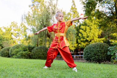 7 yaşında, kırmızı spor Wushu üniformalı, yaz günü parkta egzersiz yapan tatlı beyaz bir kız. Kung fu savaşçı çocuk atletinin yaşam tarzı portresi