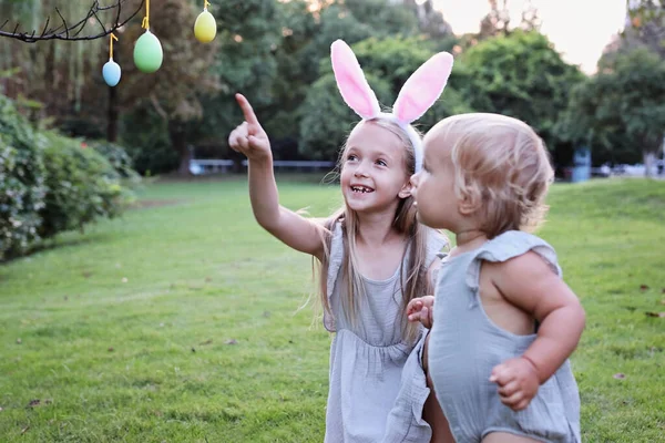 Dos niños que usan orejas de conejo cuando recogen huevos de Pascua pintados en el jardín o el parque. lindo caucásico bebé y hermana pasando tiempo juntos en patio trasero — Foto de Stock