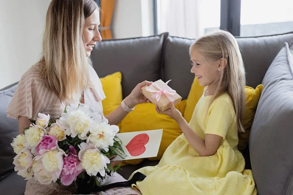 La figlia di bambino si congratula con madre e dà la carta regalo, il regalo e il mazzo di fiori a casa. Mamma e ragazza sorridono e si abbracciano sul divano. Felice festa del giorno delle madri — Foto Stock