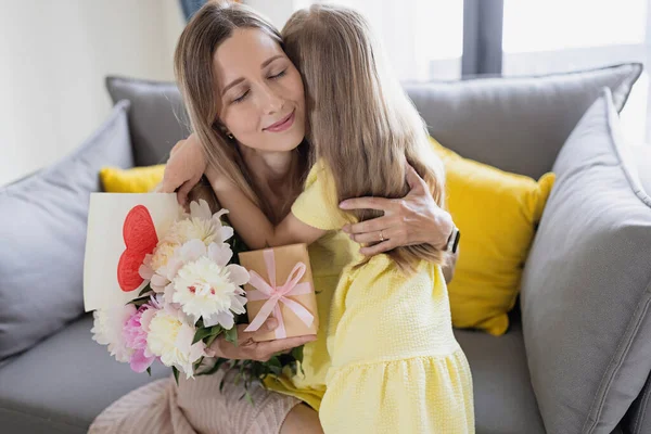 La hija del niño felicita a la mamá y regala la tarjeta regalo, el regalo y el ramo de flores en casa. Mamá y niña sonriendo y abrazándose en el sofá. Feliz celebración del día de las madres Fotos de stock