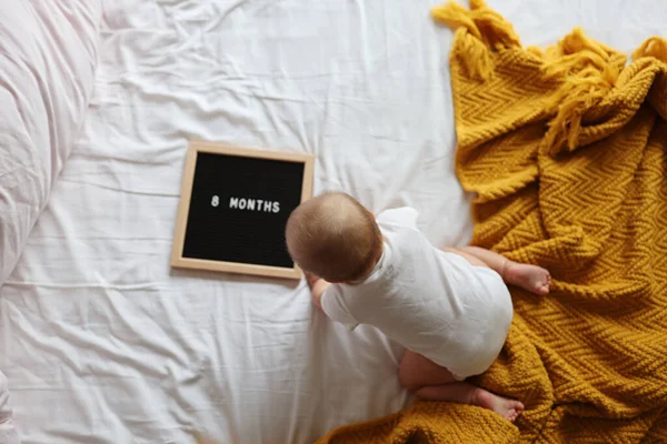 Blanke blonde baby acht maanden oud liggend op gezellig bed met gele gebreide deken thuis. Kind met witte kleding — Stockfoto