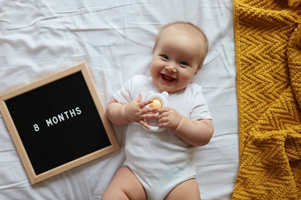 Bebé rubio caucásico de ocho meses acostado en una cama acogedora con manta de punto amarillo en casa. Niño usando ropa blanca Imagen de archivo