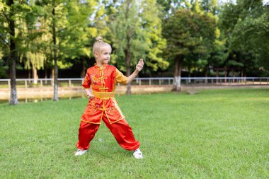 7 yaşında, kırmızı spor Wushu üniformalı, yaz günü parkta egzersiz yapan tatlı beyaz bir kız. Kung fu savaşçı çocuk atletinin yaşam tarzı portresi