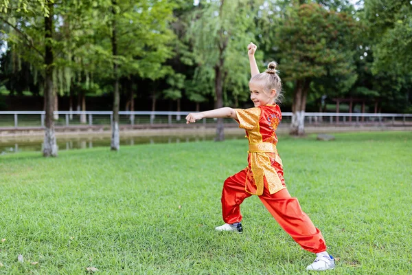 Cute little caucasian girl seven years old in red sport wushu uniform exercising in park at summer day. Lifestyle portrait of kung fu fighter child athlete — Stock Photo, Image