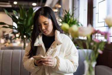 Gülümseyen Asyalı Kadın modern kafeteryada yayın yapıyor ve cep telefonu kullanıyor.