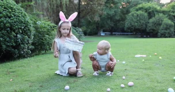 Dos niños que usan orejas de conejo cuando recogen huevos de Pascua pintados en el jardín o el parque. Lindo bebé caucásico y hermana pasar tiempo juntos en el patio trasero. Movimiento lento — Vídeo de stock