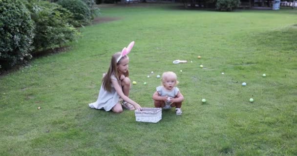 Dos niños que usan orejas de conejo cuando recogen huevos de Pascua pintados en el jardín o el parque. Lindo bebé caucásico y hermana pasar tiempo juntos en el patio trasero. Movimiento lento — Vídeo de stock