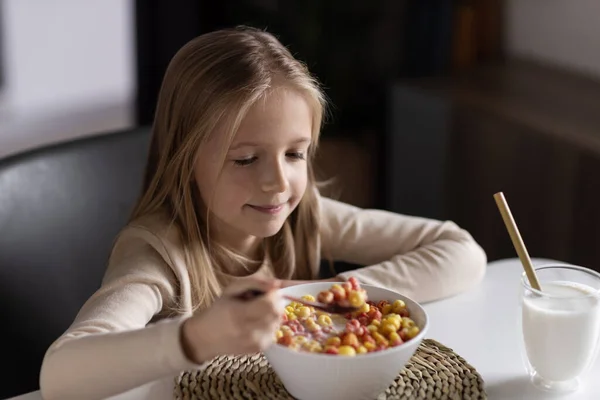 Linda niña caucásica sentada a la mesa en la cocina temprano en la mañana y preparando el desayuno con coloridos copos de maíz y leche. Niño disfrutando de la vida con comida saludable, concepto de estilo de vida saludable Fotos de stock