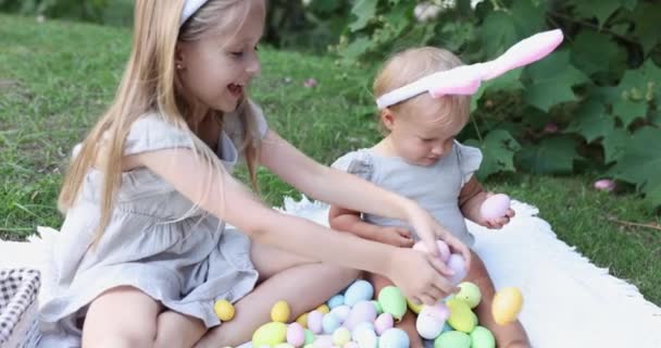 两个小孩在花园或公园捡彩绘复活节彩蛋打猎时佩戴兔耳。可爱的高加索婴儿和姐姐花时间一起在后院。慢动作 — 图库视频影像