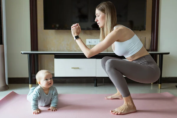 Ung mamma i sportkläder motionerar hemma med barnet. Online-utbildning under coronavirus covid-19 karantän. Håll dig i form och säker under pandemisk låsning. Sport, fitness, hälsosamt koncept Stockbild